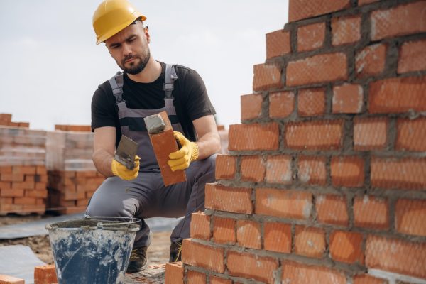 Sitting and building the foundation. Construction worker is at job with bricks outdoors.